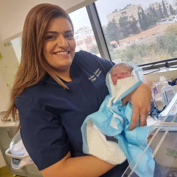 امرأة ترتدي زياً طبياً كحلياً وتبتسم، تحمل بين ذراعيها طفلاً حديث الولادة ملفوفاً ببطانية زرقاء وبيضاء بجانب نافذة.