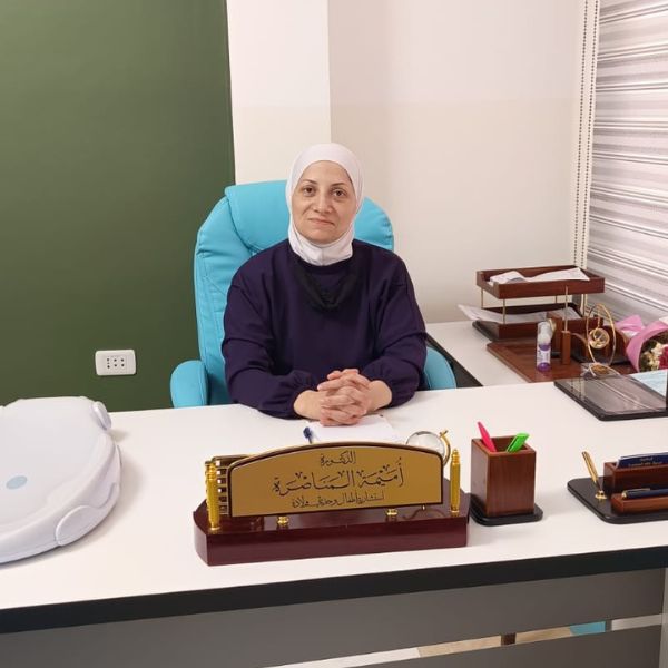 امرأة ترتدي حجاباً أبيض وقميصاً كحلياً تجلس خلف مكتب أبيض وتضع يديها متشابكتين، وعلى المكتب لافتة اسم وأدوات مكتبية وميزان أطفال.