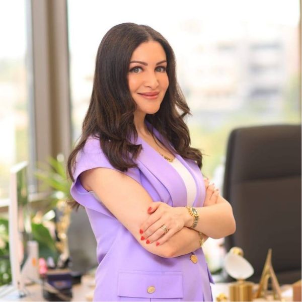امرأة ذات شعر داكن مموج ترتدي سترة بدون أكمام باللون البنفسجي، تقف مكتوفة الأيدي وتبتسم في مكتب.