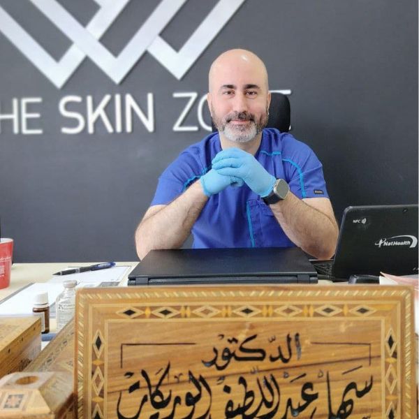 رجل أصلع بلحية يرتدي زياً طبياً أزرق وقفازات زرقاء يجلس خلف مكتب عليه حاسوب محمول ولوحة خشبية مزخرفة.