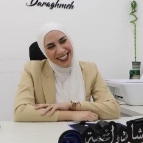 امرأة ترتدي حجاباً أبيض وسترة باللون البيج تبتسم وهي تجلس خلف مكتب.