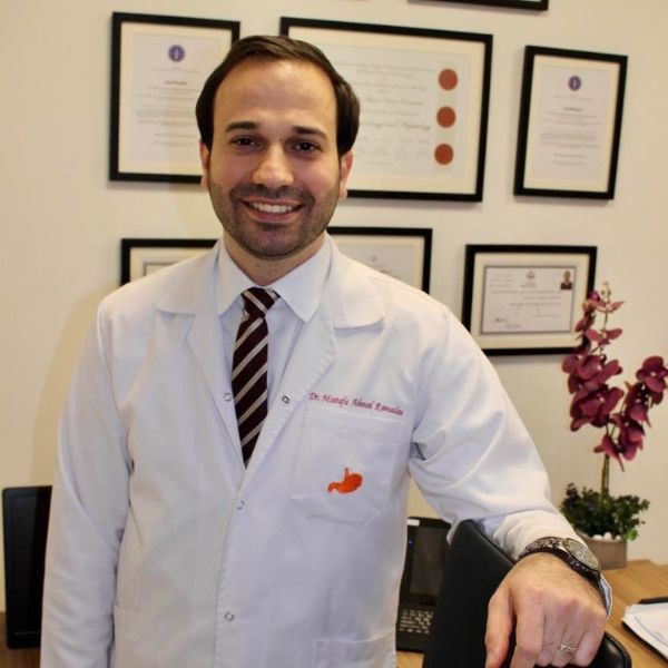 رجل يبتسم يرتدي معطفًا طبيًا أبيض وربطة عنق، وتقع خلفه شهادات مؤطرة معلقة على الجدار ونبات زينة.