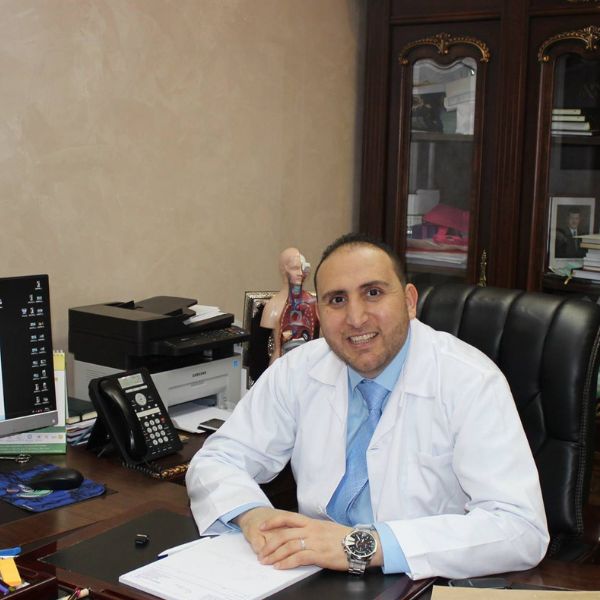 رجل يبتسم ويرتدي معطفًا طبيًا أبيض وقميصًا أزرق، يجلس خلف مكتب عليه هاتف وطابعة وأوراق، وخلفه خزانة خشبية ومجسم تشريحي.
