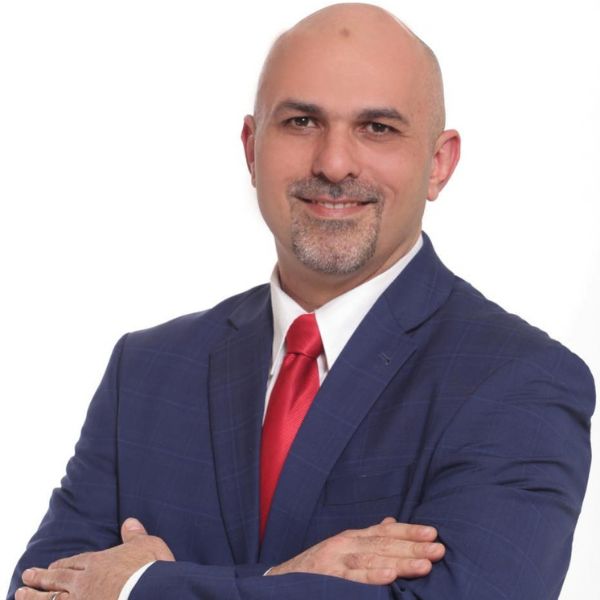 صورة شخصية لرجل أصلع يبتسم، يرتدي بدلة كحلية وقميصاً أبيض وربطة عنق حمراء، مكتف اليدين أمام خلفية بيضاء.